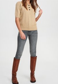 Beżowy wełniany sweter z okrągłym dekoltem i trzema ciemnymi guzikami, w połączeniu z dopasowanymi szarymi dżinsami i wysokimi brązowymi skórzanymi butami.