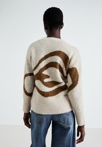 Beige strikket genser med et brunt abstrakt design på baksiden, med ribbestrikket kant og mansjetter, som gir en myk tekstur.