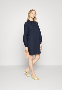 Vestido azul-marinho com botões, mangas longas e volumosas, e cintura ajustada. Combinado com sapatos de salto amarelos abertos e uma carteira rosa fofo. Textura suave.