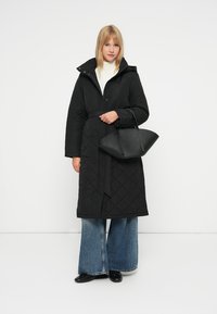 Juodas siuvinėtas ilgas paltas su kapisonu ir diržu, derinamas su per didelėmis džinsinėmis kelnėmis. Modelis laiko juodą, struktūrizuotą rankinę.