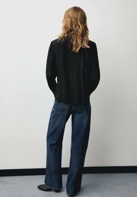 Zwarte lange mouwen shirt en losse blauwe spijkerbroek gezien vanuit de rug, met rechte pijpen en minimale details, tegen een neutrale achtergrond.