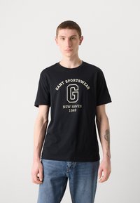 Sort bomulds t-shirt med korte ærmer, der har et hvidt og gyldent "GANT SPORTSWEAR" logo og "NEW HAVEN 1949" tekst. Casual fit.