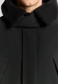 Svart kappa med en texturerad svart pälskrage, slät tyg, framsömnadsdetaljer och stora snedställda fickor på varje sida.