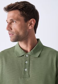 Zelena polo majica meke teksture, s ovratnikom, pločicom s četiri gumba i diskretnim uzorkom okomitih pruga.