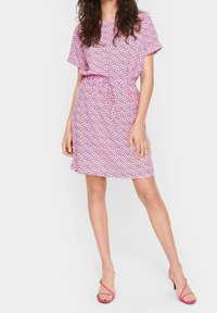 Rosa kortärmad klänning med svarta hjärtformat mönster, figursydd midja och utsvängd nederkant. Stylad med rosa sandaler med remmar.