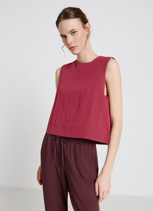 Donna con i capelli legati indossa una camicetta rossa senza maniche e pantaloni larghi color bordeaux su sfondo chiaro e semplice.