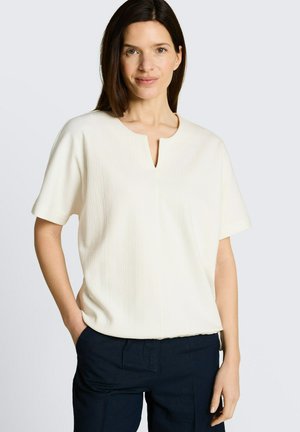 Frau mit schulterlangem dunklem Haar, die ein cremefarbenes Kurzarmoberteil mit kleinem V-förmigem Ausschnitt und dunkle Hosen trägt, Hände entspannt.