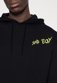 Felpa nera con cappuccio e cordini, dal tocco morbido; presenta la scritta "SAD BOY" stampata in giallo brillante sul davanti.