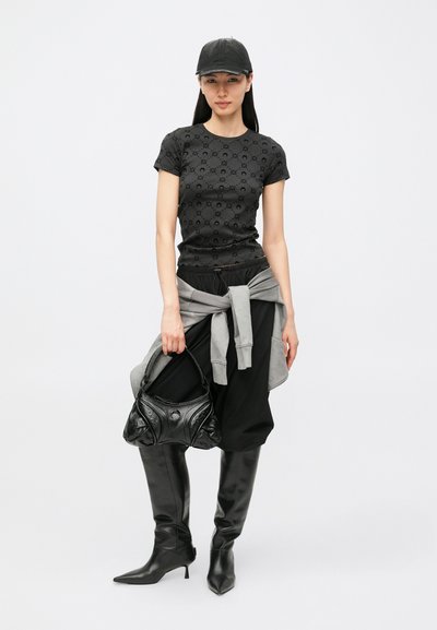 T-shirt preta com padrão, suéter cinzento amarrado à cintura, calças pretas largas, botas pretas altas de salto e boné preto.