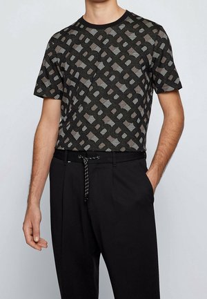 Mann trägt ein schwarz-grau geometrisch gemustertes kurzärmeliges Hemd, das in schwarze Hosen mit einem schwarz-weißen Kordelgürtel gesteckt ist.
