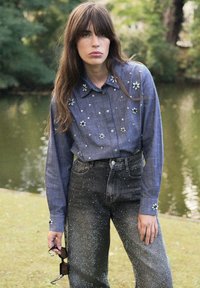 Chemise en denim à boutons avec des embellissements floraux, associée à un jean noir taille haute orné de paillettes éparpillées. Lunettes de soleil à la main.