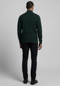 Jersey de punto acanalado en verde oscuro con cuello alto, que presenta sutiles detalles en los hombros, combinado con pantalones negros y botas negras.