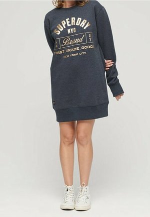 Persoon die een donkergrijze Superdry NYC lange sweaterjurk draagt met gouden letters en witte hoge sneakers, staand tegen een effen achtergrond.