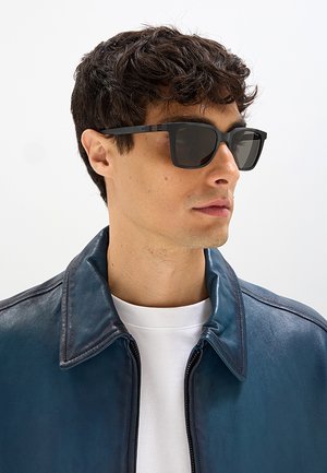 Giovane uomo con capelli corti e ricci che indossa occhiali da sole neri rettangolari, una giacca di pelle blu e una camicia bianca, guarda di lato.