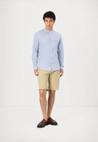Camicia azzurro chiaro con un finish testurizzato, pantaloni corti khaki, e mocassini marroni. La camicia presenta una tasca sul petto e un dettaglio del logo.