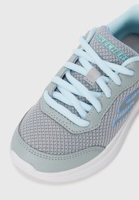 Grauer Sportschuh mit Mesh-Obermaterial, hellblauen Schnürsenkeln und blauem Logo-Akzent. Runde Zehenpartie und weiße Gummisohle.