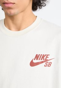 Krémové bavlnené tričko s červeným logom Nike SB na ľavej strane hrudníka. Klasický strih s hladkou textúrou.
