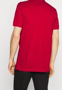 Punainen lyhythihainen polo-paita pehmeällä tekstuurilla, klassinen kaulus ja sivuaukot helmassa. Sen kanssa on käytetty mustia housuja.