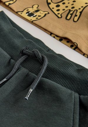 Mørkegrønne sweatpants med et struktureret ribbet livrem og grå snore. Et mønstret tan stof med leopardmotiver er synligt ovenfor.