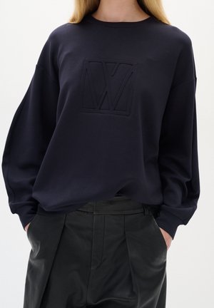Person trägt einen marineblauen Pullover mit einem geprägten "VW"-Logo und schwarze, hoch taillierte Lederhosen, Hände in den Taschen, neutraler Hintergrund.