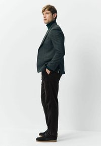 Man in dark gray blazer, green turtleneck, dark brown corduroy pants, and black shoes standing with hands in pockets against white background.
