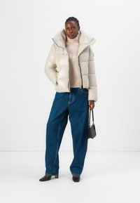 Pufferjack in lichtbeige met een hoge kraag, ritssluiting en een gestructureerde afwerking. Gecombineerd met losse blauwe denim broeken en zwarte schoenen.