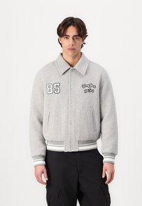 Giacca bomber in lana grigia con chiusura a bottoni, polsini e orlo a costine. Presenta dettagli ricamati: "85" e "Tommy Jeans New York."
