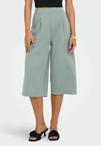 Spodnie o szerokich nogawkach z miękkiego, stonowanego zielonego materiału. Posiadają wysoką talię i schludne plisowania z przodu. Połączone z czarnymi mule na obcasie z kwiatowymi akcentami.