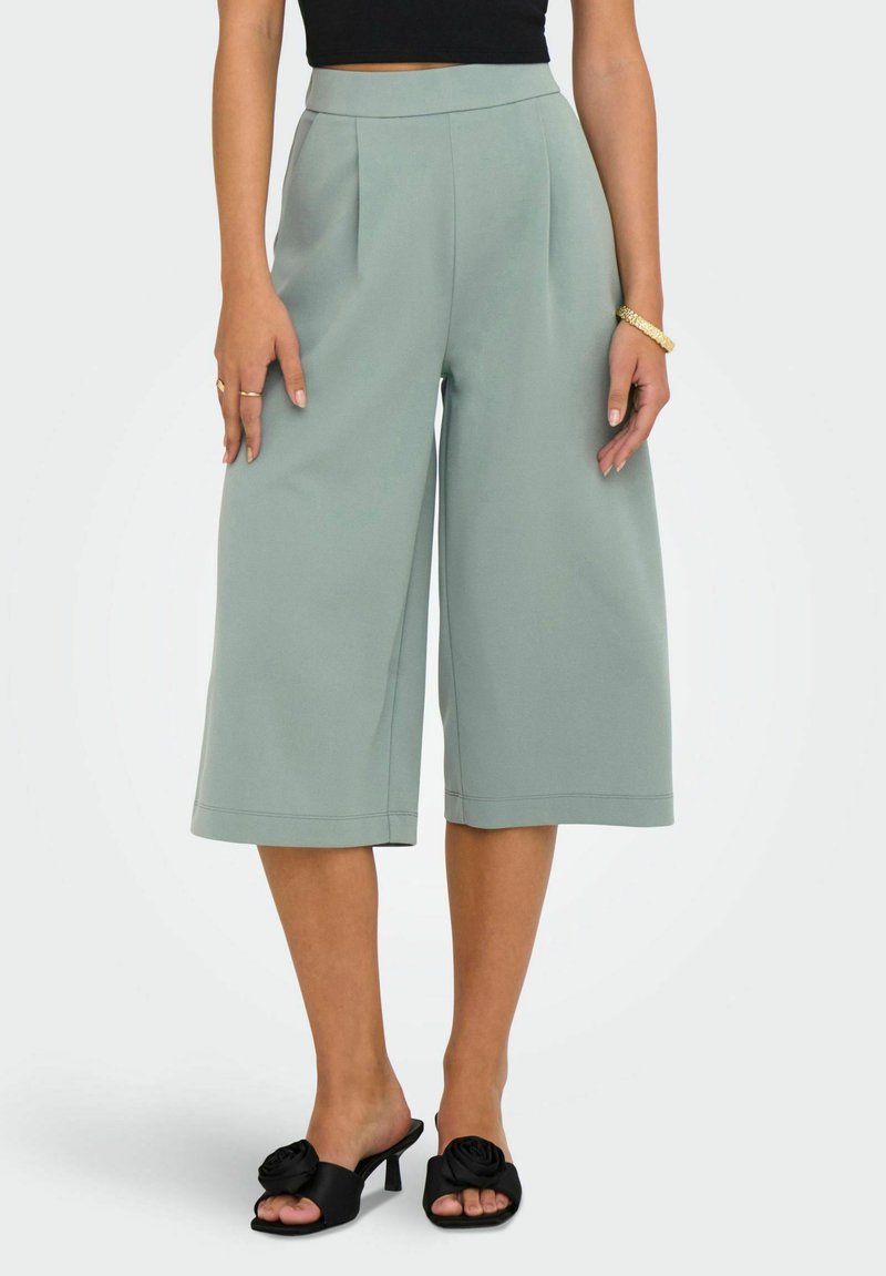 Spodnie o szerokich nogawkach z miękkiego, stonowanego zielonego materiału. Posiadają wysoką talię i schludne plisowania z przodu. Połączone z czarnymi mule na obcasie z kwiatowymi akcentami.
