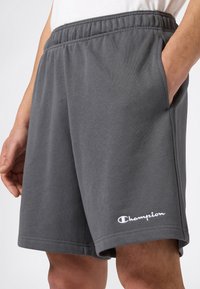 Uomo che indossa pantaloni corti Champion grigi scuri con cintura elasticizzata e tasche laterali, con una mano in tasca, su uno sfondo bianco.