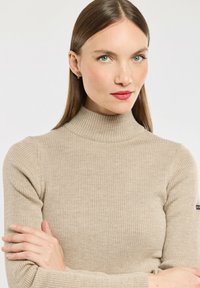 Femme aux longs cheveux bruns portant un pull à col roulé beige à côtes, les bras croisés, avec une expression neutre devant un fond uni.