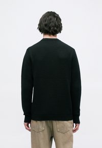 Μαύρο υφασμάτινο πλεκτό πουλόβερ με στρογγυλή λαιμόκοψη και ριχτά μανίκια, φορεμένο πάνω από ανοιχτά καφέ παντελόνια, κοιτώντας από πίσω.