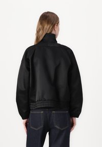 Black bomber jacket with a high collar and elasticated cuffs. Smooth, shiny fabric; ribbed hem; worn over blue denim jeans.