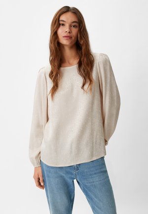 Light beige blouse with shimmering, textured fabric featuring a repeating logo pattern. Long sleeves gather at the wrist, rounded hem design.