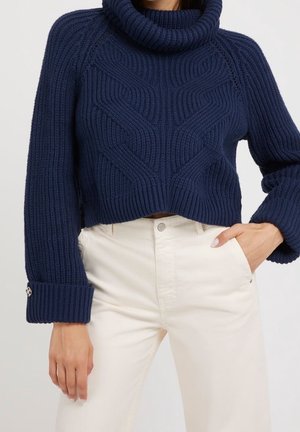 Woman wearing a navy blue chunky knit turtleneck sweater with cable patterns and cream high-waisted pants, hand in pocket.
