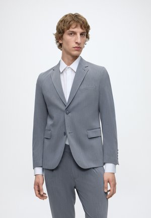 Jeune homme aux cheveux bouclés châtain clair portant un costume gris clair et une chemise blanche sur un fond blanc uni.