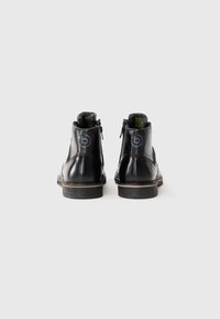 Botas de cuero negras hasta el tobillo con cremalleras laterales, con el logo de la marca visible en el tacón y la suela, fotografiadas desde la parte trasera sobre un fondo blanco.