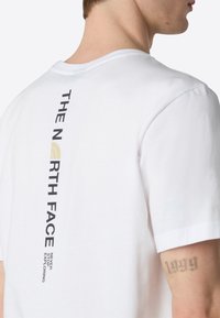 Hvid bomulds t-shirt med korte ærmer, der har et vertikalt sort og guld logo tekstdesign på ryggen, glat tekstur, afslappet pasform.