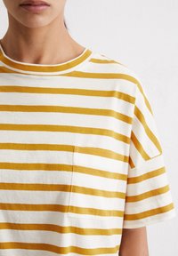 Geel en witte gestreepte katoenen t-shirt met een ronde hals en korte mouwen. Bevat een borstzak aan de linkerkant.