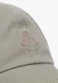 Beige baseball cap made of textured fabric, featuring an embroidered skull and crossbones design in light brown on the front.