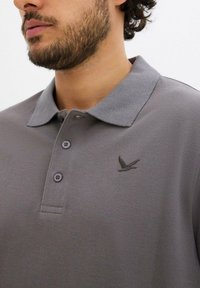 Maglietta polo grigia realizzata in tessuto testurizzato, con colletto classico, tre bottoni e un sottile logo di un uccello ricamato sul petto.