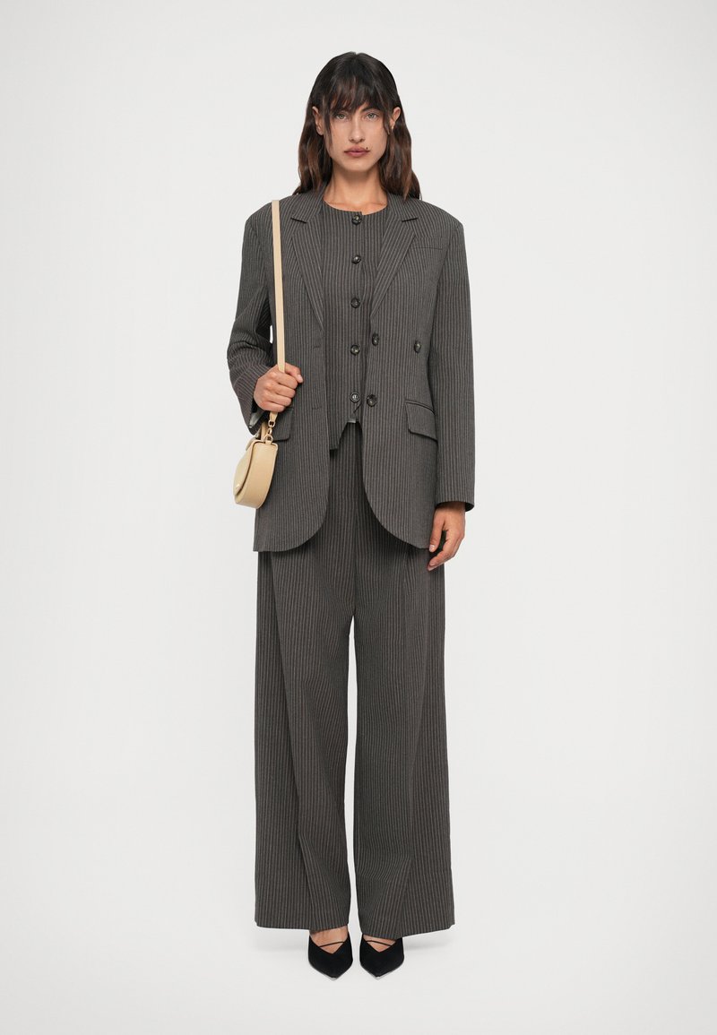 Pinstripe suit in dark gray, featuring a tailored blazer with double buttons, loose-fit trousers, and black pointed toe heels. A small beige crossbody bag is held.