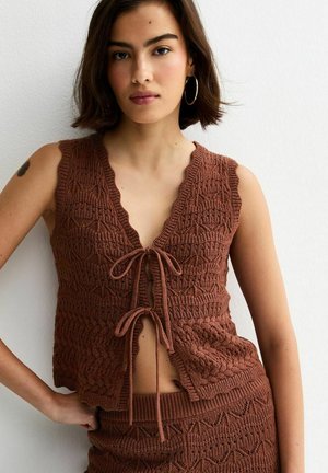 Giovane donna con top marrone lavorato a maglia senza maniche, annodato davanti con due fiocchi, e gonna abbinata, in piedi contro una parete bianca.