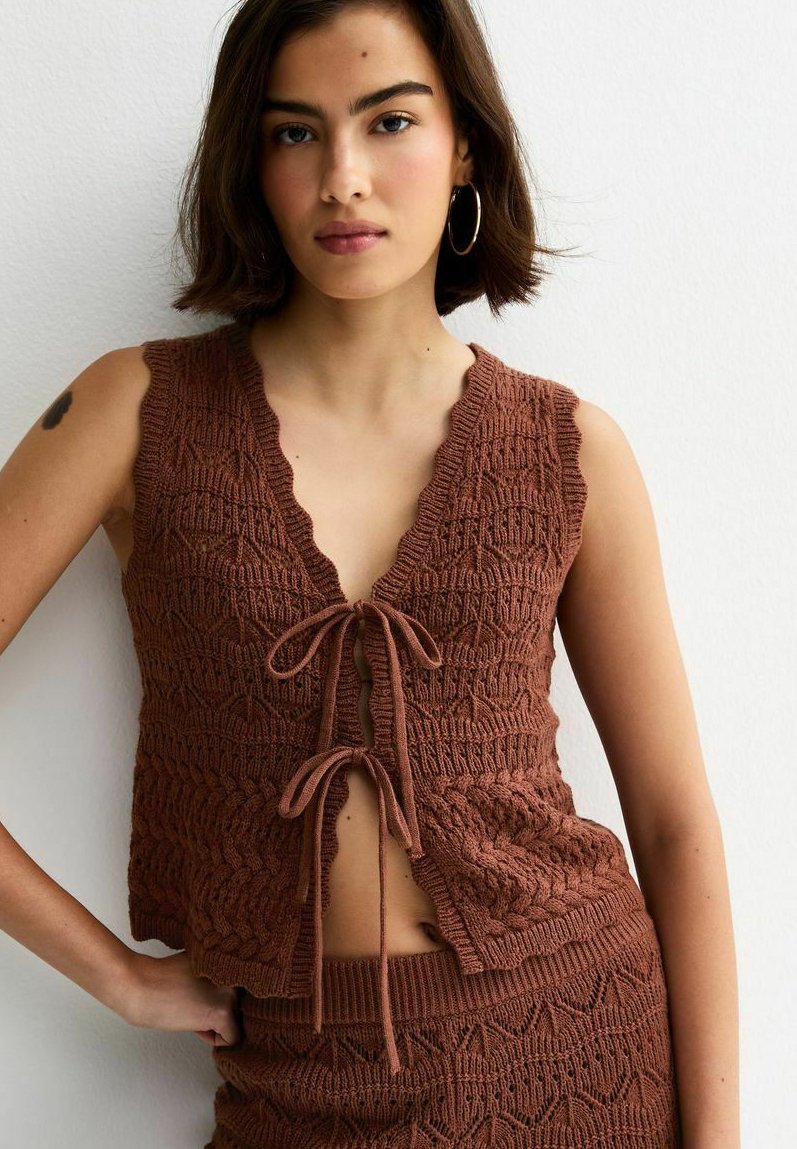 Młoda kobieta w brązowym, bezrękawnikowym, dzianinowym topie zawiązanym na dwa kokardy z przodu oraz w dopasowanej spódnicy, stojąca na tle białej ściany.