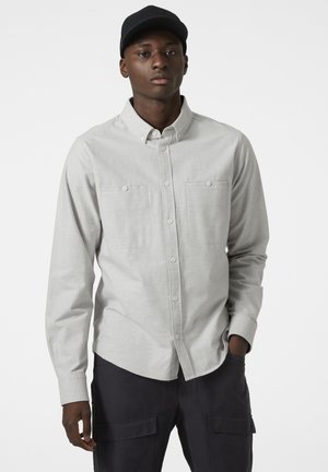 Camisa de botones gris claro con dos bolsillos en el pecho, mangas largas y un cuello clásico. Combinada con pantalones cargo oscuros.