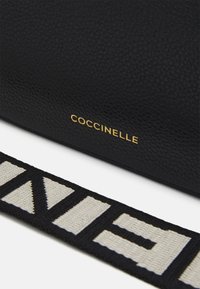 Černá texturovaná kožená kabelka se zlatým logem "Coccinelle". Má popruh z vzorované tkaniny v černých a béžových tónech s geometrickými motivy.