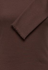 Gros plan d’un tissu tricoté côtelé marron avec une manche longue présentant un ourlet à bord ondulé type laitue.