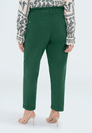 Frau trägt hoch taillierte grüne Hose mit Gesäßtaschen, kombiniert mit einer langärmeligen Bluse mit Paisley-Muster und beige Sandalen mit hohen Absätzen.