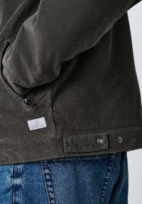 Primer plano de una chaqueta de pana marrón oscuro con un bolsillo con cremallera y etiqueta, llevada sobre unos jeans de mezclilla azul.
