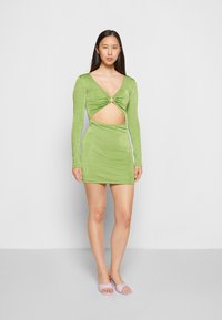 Vestido verde de mangas longas com decote em V profundo, detalhe torcido no busto, cintura recortada e uma textura suave. Acompanhado por sandálias slide em rosa pastel.